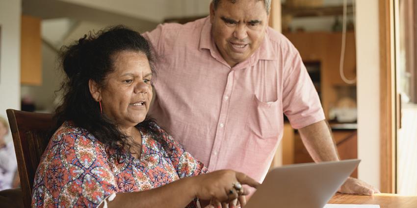 This image shows two people working together at a computer. The person on the left, wearing a colorful, patterned blouse, is seated and pointing at the laptop screen, engaged in explaining something. The person on the right, dressed in a pink shirt, is standing slightly behind, looking intently at the screen, appearing to listen or observe carefully. 