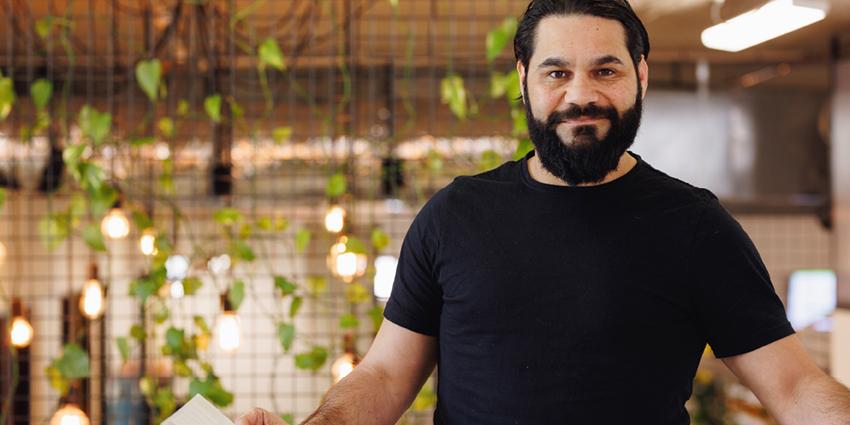This image shows a man standing in a casual, modern indoor setting. He has a beard and is wearing a black T-shirt, smiling slightly as he looks at the camera. He is holding a book or notebook in one hand, suggesting he might be presenting or explaining something. The background features a decorative wall with hanging plants and visible string lights, creating a warm and inviting atmosphere, typical of a relaxed workspace or creative environment.