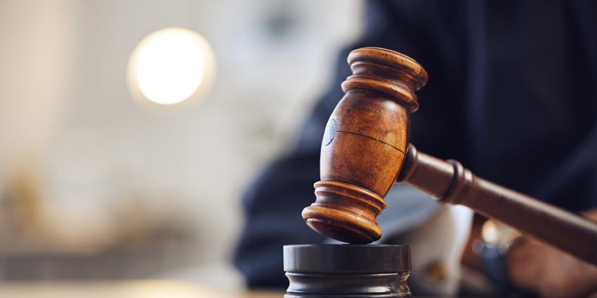 This image features a close-up of a wooden gavel resting on a sound block, commonly associated with judicial or legal proceedings. The gavel, held by a hand in the background, symbolizes justice and authority. The setting appears to be a courtroom or formal legal environment, with soft, focused lighting highlighting the gavel, making it the central focus.