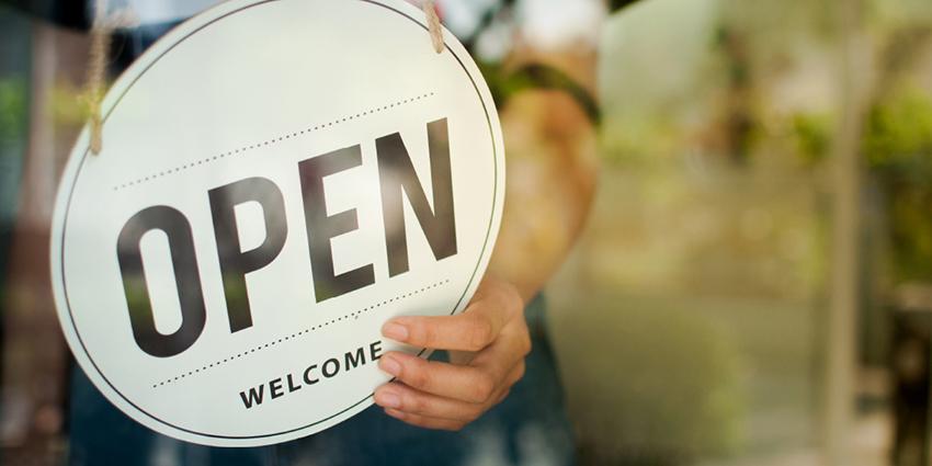 This image shows a close-up of a hand holding a circular "OPEN" sign with "WELCOME" written beneath it. The sign is being displayed in front of a glass door or window, indicating that a business is open and ready to welcome customers. The background is slightly blurred, with hints of greenery visible, suggesting an inviting and friendly atmosphere.