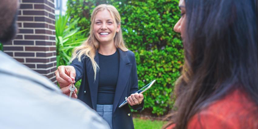 This image shows a smiling real estate agent handing over a set of keys to a couple, symbolizing the successful completion of a property transaction. The agent, dressed professionally in a blazer, holds a tablet in one hand while extending the keys with the other, indicating a friendly and celebratory handover. 