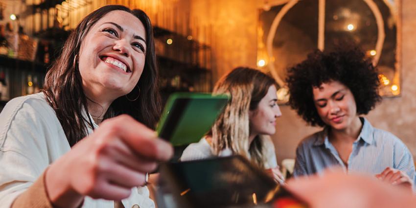 This image shows a joyful woman holding out a green credit card, likely making a payment. She is smiling brightly, creating a warm and friendly atmosphere. Behind her, two other women are seated, engaged in conversation, adding a social and relaxed vibe to the scene. 