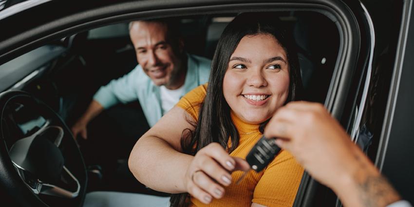 This image shows a cheerful young woman sitting in the driver’s seat of a car, smiling as she reaches out to receive a set of car keys from someone outside the vehicle. A man seated beside her in the passenger seat is also smiling, suggesting a happy moment, possibly related to purchasing or renting the car.