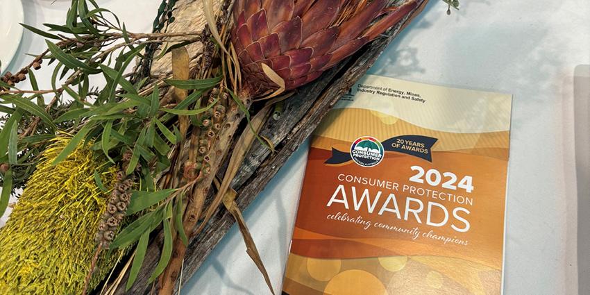 This image features a brochure for the "2024 Consumer Protection Awards" with the subtitle "celebrating community champions." The brochure is displayed on a table with a decorative arrangement of native Australian flora, including branches and dried flowers.