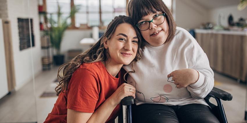 This warm and uplifting image captures two women in a cozy indoor setting. One woman, wearing a red shirt, is sitting closely beside another woman in a wheelchair, who has glasses and a joyful expression. They are smiling and appear connected, conveying a sense of friendship, support, and inclusion.