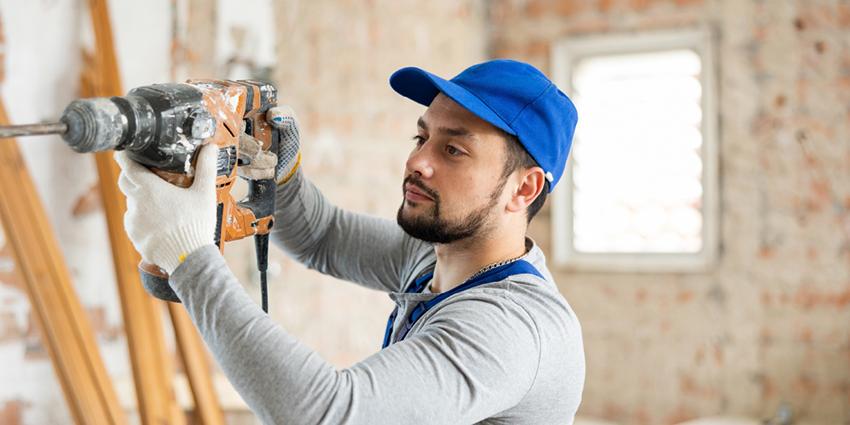 This image features a tradesperson in a work setting, wearing a blue cap, gloves, and gray shirt, operating a heavy-duty drill.