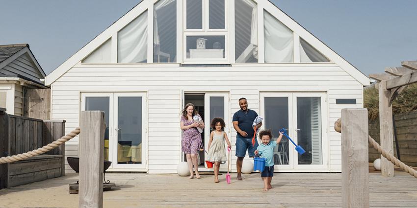 This warm image shows a family exiting a beach house or holiday home. The family, consisting of adults and children holding beach toys, appears happy and ready for a day out.
