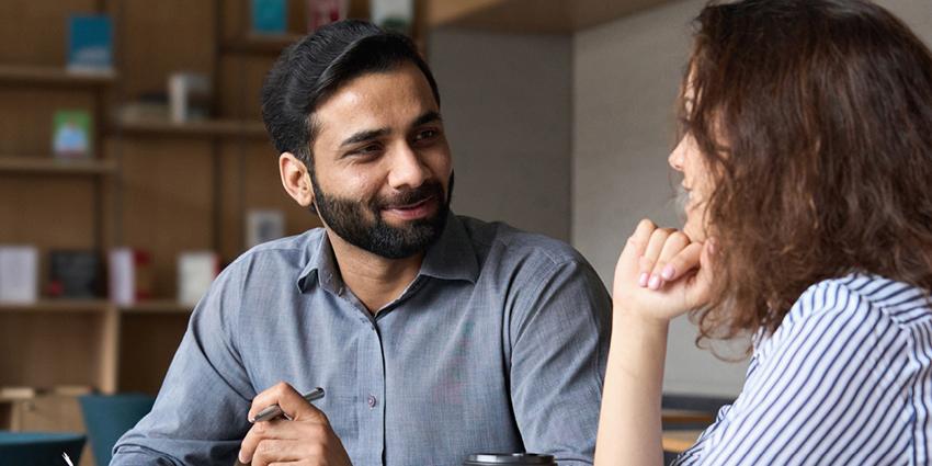 This image shows two individuals engaged in a friendly conversation, likely in a professional setting. Both are seated, and the man appears attentive as the woman speaks.