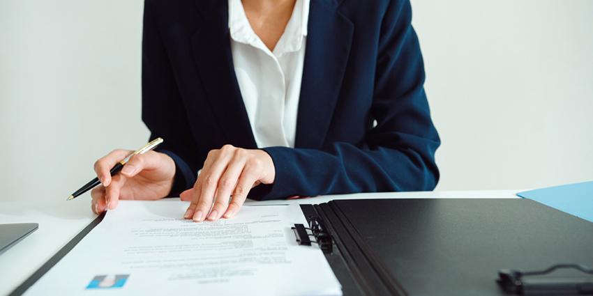 A close-up of a professional at a desk, reviewing or signing documents. The individual is dressed in a business suit, and the setting is formal, with neatly arranged paperwork.