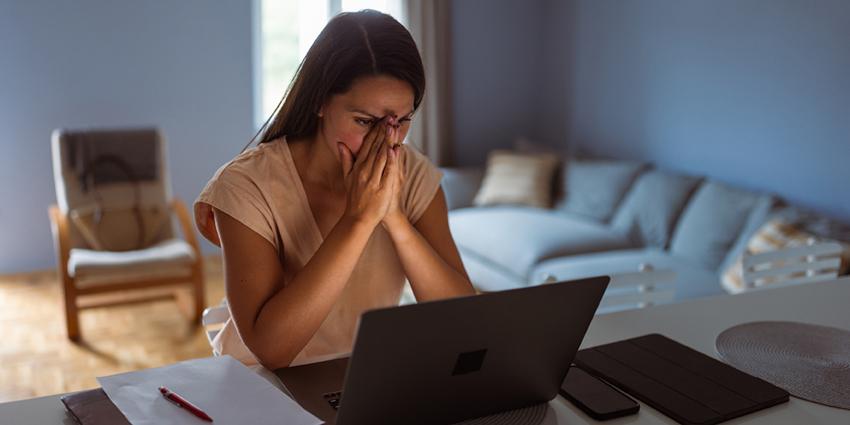 A woman looking stressed or frustrated, seated at a table with her hands over her face, looking at a laptop screen.