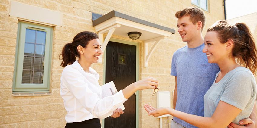 A smiling real estate agent handing keys to a young couple standing outside a home. The couple looks happy and excited, symbolizing the accomplishment of buying or renting a new home.