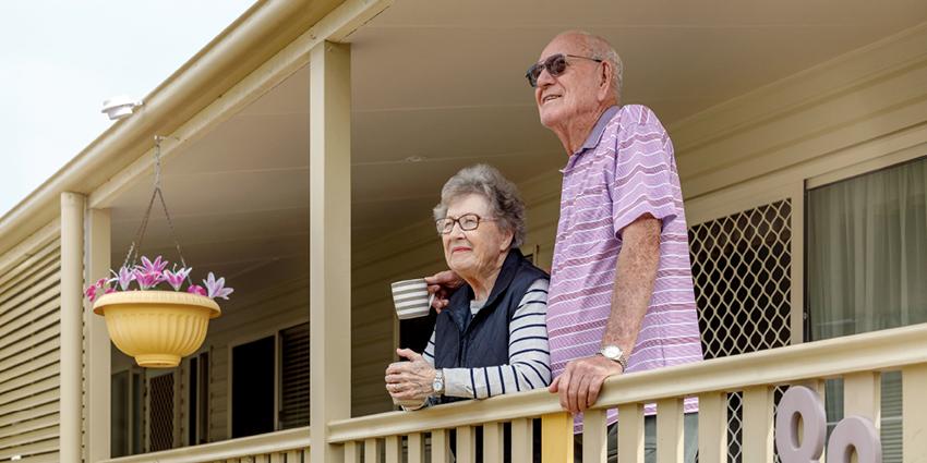 An elderly couple standing on the porch of their home, looking content. They are dressed casually, and the scene is warm and relaxed.