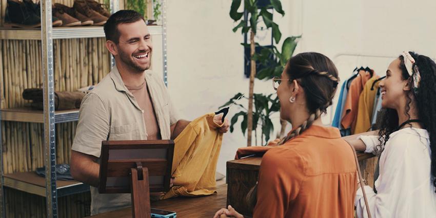 This image shows a friendly interaction at a retail counter. A smiling salesperson, dressed casually, is assisting two female customers who are also smiling and engaged in the conversation.