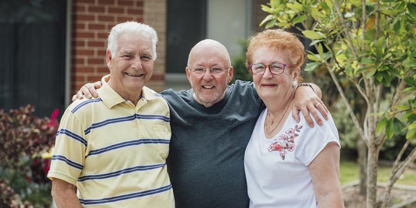 This image captures three elderly friends standing together outside, smiling warmly. The trio is posing with their arms around each other, conveying a sense of companionship and community.